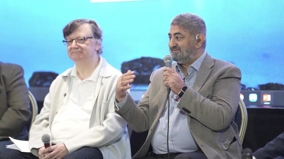 Still from the AI in Action video: a speaker holding a microphone on a panel